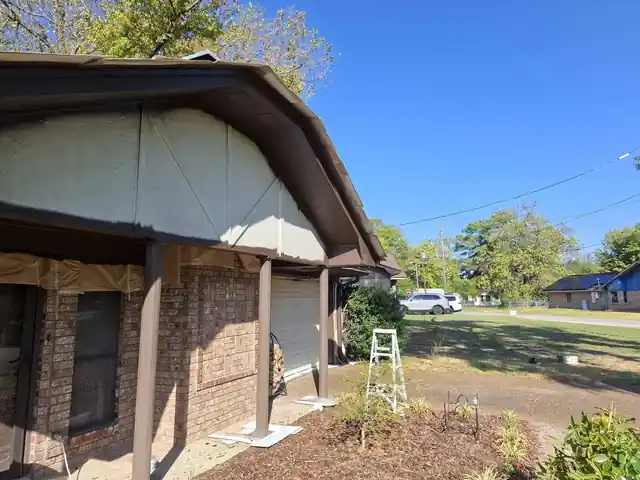 Brick ranch mid-repaint: brown masking paper over every window, cream gable panels freshly coated, a ladder against the eave