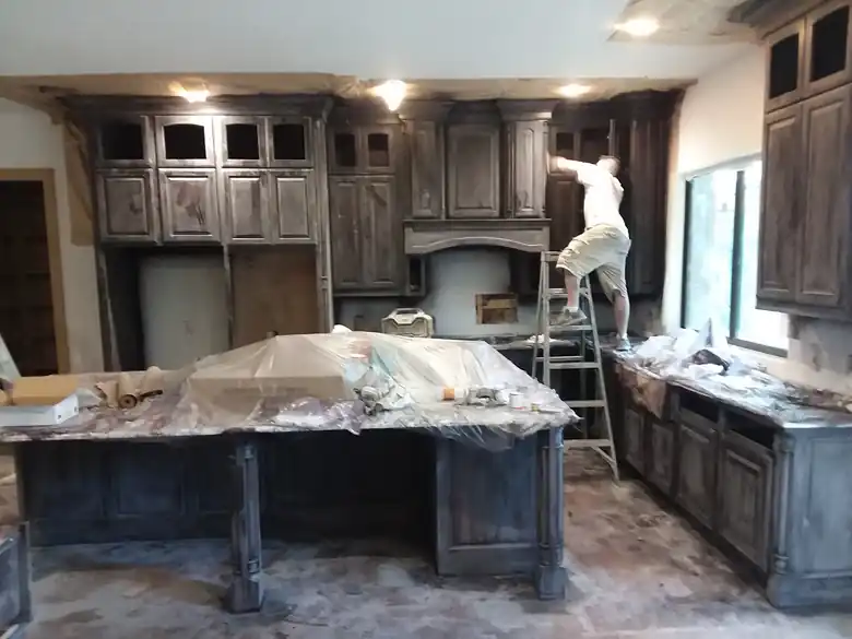 Kitchen cabinets being sprayed dark under poly, a man on a ladder with his back to the camera