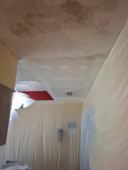 A ceiling partway through its skim coat: a smooth patched section beside the older stained section, walls sheeted in plastic